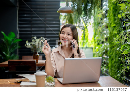 An attractive Asian businesswoman is talking on the phone while working remotely from a coffee shop. 116137957