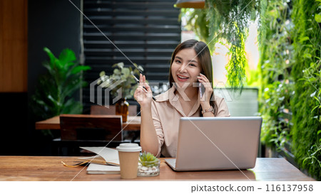 An attractive Asian businesswoman is talking on the phone while working remotely from a coffee shop. An attractive Asian businesswoman is talking on the phone while working remotely from a coffee shop. 116137958
