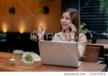 A confident Asian businesswoman working remotely from a cafe, talking on the phone with her client. A confident Asian businesswoman working remotely from a cafe, talking on the phone with her client. 116137959