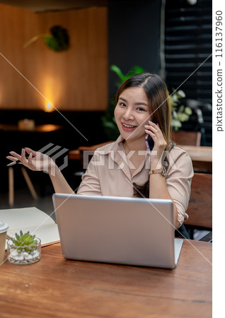 A confident Asian businesswoman working remotely from a cafe, talking on the phone with her client. A confident Asian businesswoman working remotely from a cafe, talking on the phone with her client. 116137960