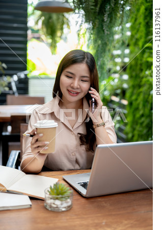 A businesswoman is talking on the phone and having a coffee while working remotely from a cafe. A businesswoman is talking on the phone and having a coffee while working remotely from a cafe. 116137961