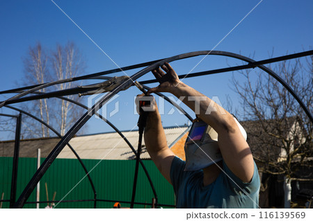 A man makes a greenhouse in the garden by welding it from pipes. A male welder wears a protective eye mask and works in the garden. A man puts up a greenhouse, welds pipes and erects a frame. A man makes a greenhouse in the garden by welding it from pipes. A male welder wears a protective eye mask and works in the garden. A man puts up a greenhouse, welds pipes and erects a frame. 116139569