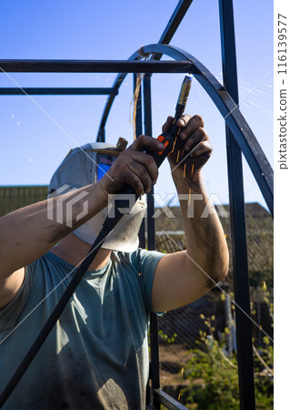 Vertical. A man welds metal in the garden wearing a protective mask in summer. A man works in his garden welding a greenhouse for seedlings. Concept of welding work in the garden. 116139577
