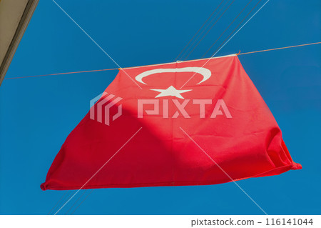 Turkey's red flag with a white crescent moon and a five-pointed star hangs on a rope between houses against a clear blue sky. Symbols of statehood, patriotism, signs, symbolism. Turkey's red flag with a white crescent moon and a five-pointed star hangs on a rope between houses against a clear blue sky. Symbols of statehood, patriotism, signs, symbolism. 116141044