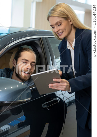 Blonde sale assistant showing something on a tablet to a customer in a dealership 116144234
