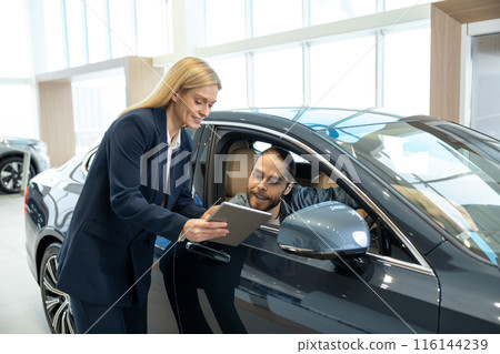 Blonde sale assistant showing something on a tablet to a customer in a dealership 116144239