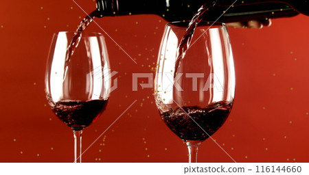 Red wine pouring into two glasses against red background, sparkling droplets visible 116144660