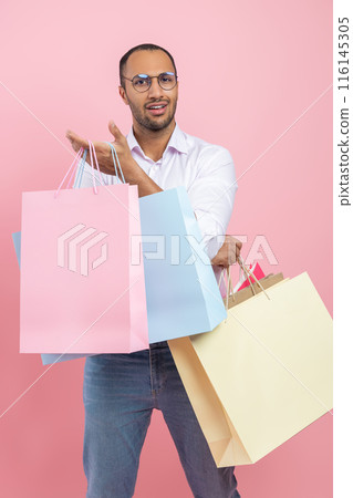Uncertain African American man wearing shirt and glasses holding shopping bags standing with questioning expression questioning expression 116145305