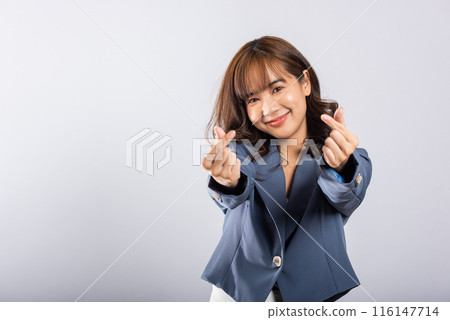 Confident business woman stands with a smile, creating a mini heart sign with her finger on a white background. Asian portrait of a beautiful young female sending love for Valentine's Day, studio shot Confident business woman stands with a smile, creating a mini heart sign with her finger on a white background. Asian portrait of a beautiful young female sending love for Valentine's Day, studio shot 116147714