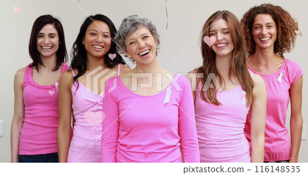 Image of pink hearts floating over diverse group of smiling women in pink with cancer ribbons on 116148535