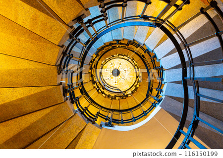 A view from the bottom of a winding staircase, looking up toward a chandelier. The staircase features a wooden handrail and metal railings. The surrounding walls are a warm, pale yellow color. 116150199
