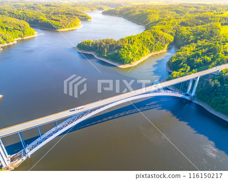 An aerial view of the Zdakov Bridge spanning the Vltava River in the Czech Republic. The bridge arches gracefully over the water, surrounded by lush green forests. 116150217