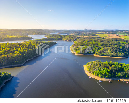 Aerial view of Orlik Water Reservoir on the Vltava River in Czechia, surrounded by forests and hills. Calm waters reflect blue sky, creating serene and idyllic landscape. 116150219