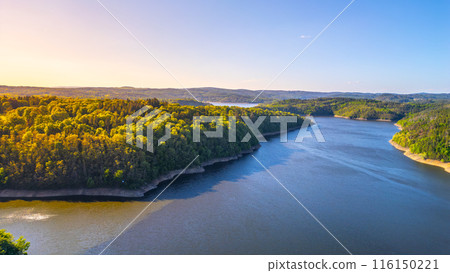 Aerial view of Orlik Water Reservoir on the Vltava River in Czechia, surrounded by forests and hills. Calm waters reflect blue sky, creating serene and idyllic landscape. 116150221