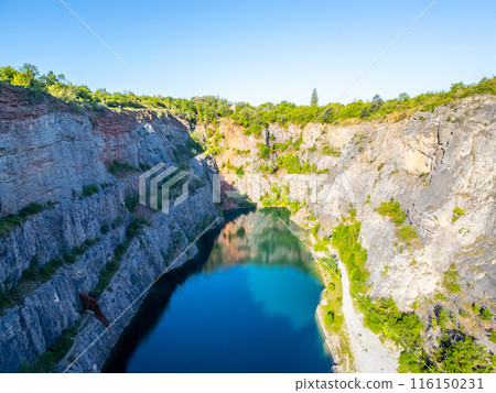 Elevated perspective of the abandoned Velka Amerika limestone quarry, featuring towering cliffs, sparse vegetation, and a reflective pond at sunset. Morina, Czechia 116150231