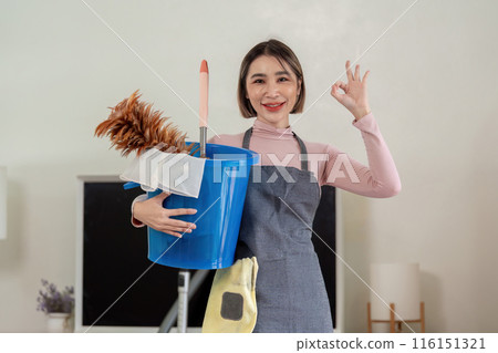 Cheerful Housekeeper Holding Cleaning Supplies and Giving OK Sign in Modern Home Interior Cheerful Housekeeper Holding Cleaning Supplies and Giving OK Sign in Modern Home Interior 116151321