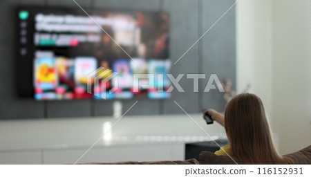 Girl selects tv channels with remote control at living room on sofa. Child controls TV using a modern remote control. 116152931