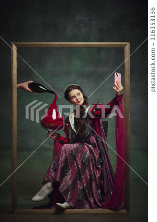 Portrait of young medieval royal person, woman sitting inside golden frame, with giant wine glass and taking selfie with mobile phone against vintage green background. 116153136
