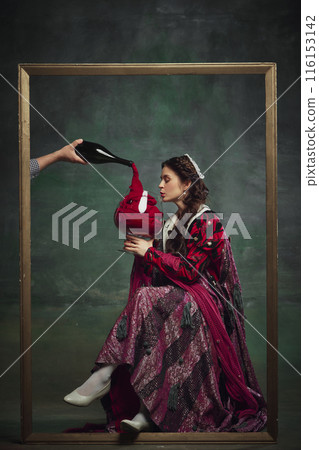 Portrait of young medieval royal person, woman sitting inside golden frame with giant wine glass against vintage green background. 116153142