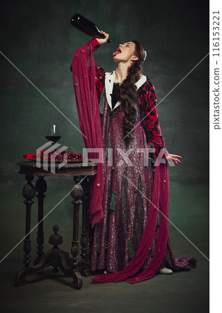 Addiction to alcohol. Young elegant woman, royal person shaking empty wine bottle on her mouth against dark green background. Addiction to alcohol. Young elegant woman, royal person shaking empty wine bottle on her mouth against dark green background. 116153221