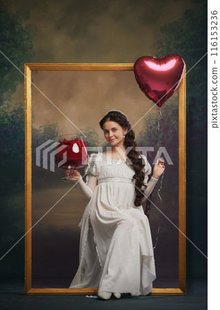 Portrait of beautiful young woman in tender white dress, sitting inside golden picture frame with heart-shaped balloon and giant wine glass. Valentine's day 116153236