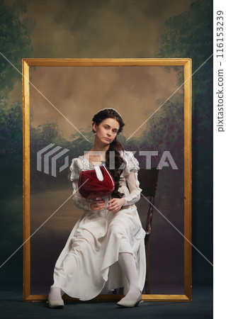 Young beautiful woman in white dress sitting into golden picture frame with giant wine glass and sad face against vintage background Young beautiful woman in white dress sitting into golden picture frame with giant wine glass and sad face against vintage background 116153239