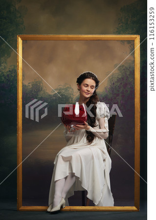 Humorous advertisements for wine brand. Beautiful young woman, medieval princess sitting inside picture frame with giant glass of fabric wine. Humorous advertisements for wine brand. Beautiful young woman, medieval princess sitting inside picture frame with giant glass of fabric wine. 116153259