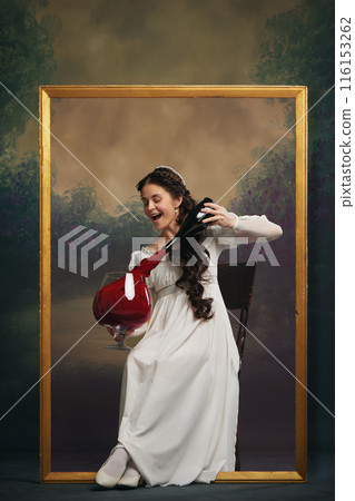 Restaurant poster promotion. Beautiful young woman in elegant white dress sitting in golden picture frame, putting wine made of fabric into giant glass 116153262