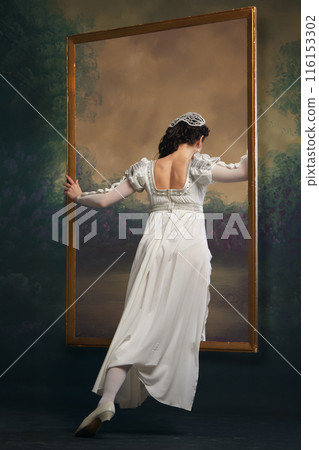 Looking into past. Young medieval woman in white dress looking inside picture frame against vintage background. Window to another era Looking into past. Young medieval woman in white dress looking inside picture frame against vintage background. Window to another era 116153302