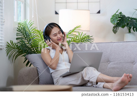 Young Woman Relax on Sofa with Headphones and Laptop, Enjoying Music in Modern Cozy Living Room Young Woman Relax on Sofa with Headphones and Laptop, Enjoying Music in Modern Cozy Living Room 116153413