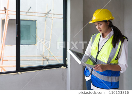 Young asian woman in a safety vest and hardhat holding solar cell panel stand next to the window. Work environment of engineer at the construction site. 116153816