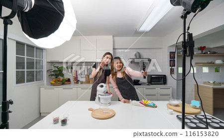 Two asian women in an apron, smiling and cooking in a kitchen with a smartphone on a selfie stick  and lighting equipment for video and photo shoot, recording vlog video for social blogger. 116153820