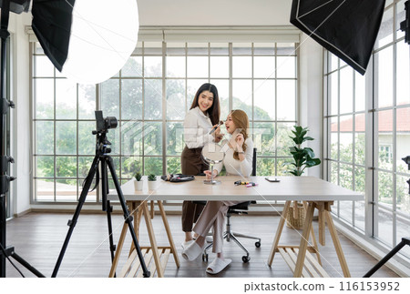 Two asian women creating content for social media. They are using a studio setup that include a camera mounted on a tripod, lighting equipment and various makeup product on the table. 116153952