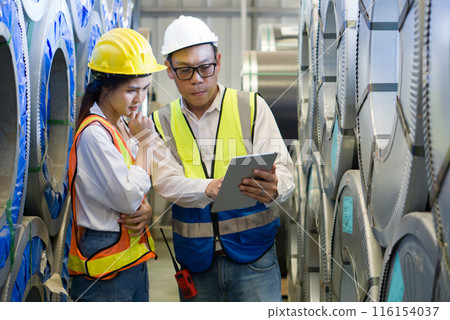 Senior engineer manager trains new employee within the metal sheet factory. Everyone wear safety vest and hardhat. Steel sheet roll stack are in the working area. 116154037