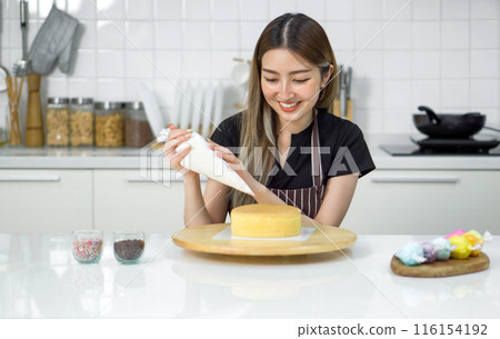 Young woman in an apron using a piping bag, pipe the icing onto the cake, creating a decorative pattern on the top of a round, light-colored cake that's placed on a wooden turntable or cake stand. Young woman in an apron using a piping bag, pipe the icing onto the cake, creating a decorative pattern on the top of a round, light-colored cake that's placed on a wooden turntable or cake stand. 116154192