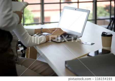 A person typing on white screen laptop computer in an office with coffee, credit card and file folder on the desk. There was a work supervisor standing behind her holding a notebook. A person typing on white screen laptop computer in an office with coffee, credit card and file folder on the desk. There was a work supervisor standing behind her holding a notebook. 116154193