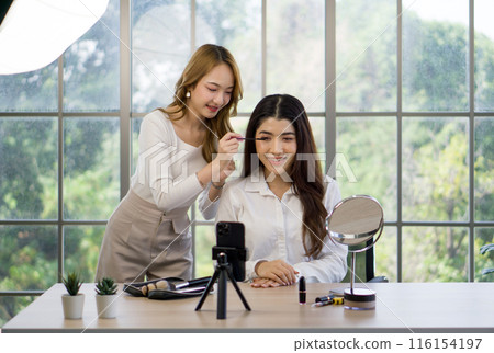Young asian makeup artist applying mascara to the sitting woman, the subject of a beauty tutorial or makeover session, given the presence of a smartphone on a tripod presumably capturing the process. 116154197