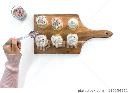 Closeup woman hand holding spoon with multicolor frosting, decorating cupcake on a wooden board, adding a colorful touch to the cupcakes' presentation. Top view 116154311