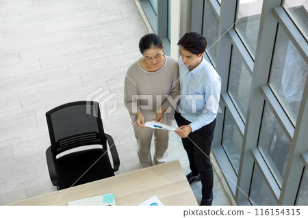 Two people in an office look at a paper with graphs. They stand next to a large window discussing it. Two people in an office look at a paper with graphs. They stand next to a large window discussing it. 116154315