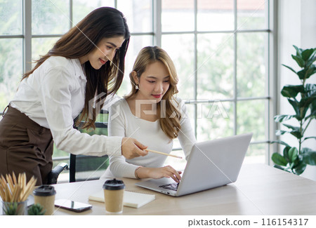 Two asian women working on laptop computer with a coffee cup, mobile phone and notebook at a desk. The background is a glass wall, overlooking the scenery of green nature Two asian women working on laptop computer with a coffee cup, mobile phone and notebook at a desk. The background is a glass wall, overlooking the scenery of green nature 116154317