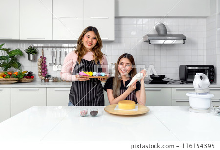 Two young women in apron make dessert in the kitchen. One people using a piping bag, pipe the icing onto the cake. The other holding wooden tray with a handful of colorful baking tools or decoration. 116154395