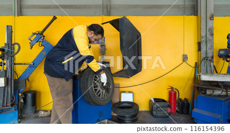 A technician in uniform and glove at an automotive service center working with a wheel balance machine. There are various garage equipment and tools in the automotive maintenance and repair sector. 116154396