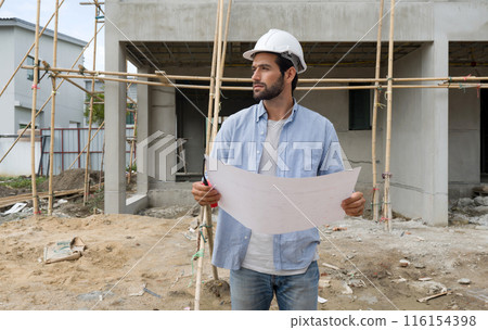 Young engineer in a construction helmet holding work calendar  while looking at the progress of real estate projects. The construction site has scaffolding made of wood. 116154398