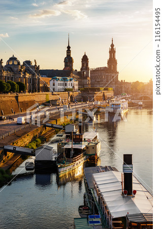 Scenic warm sunset beautiful Dresden city skyline at Elbe River with many steam tourist ship vessel moored at bank. Saxony Germany capital cityscape dusk vertical view with old Europe architecture 116154495