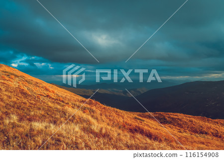 A scenic landscape view of a mountain range with a dramatic sky. The image features a foreground of rolling hills with dry grass and a distant mountain range covered in clouds. The sky is a vibrant 116154908