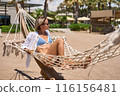 Enjoying summer vacation. Young happy woman relaxing while lying in the hammock on the sandy beach 116156481