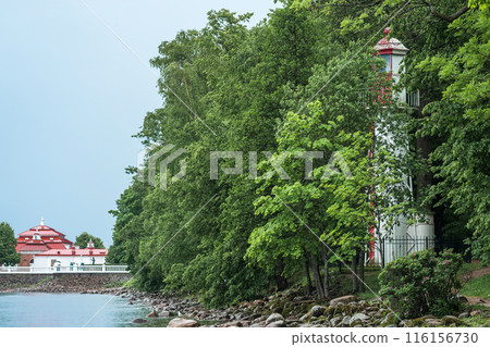 old lighthouse on the seashore among the trees against the backdrop of ancient buildings and embankment 116156730