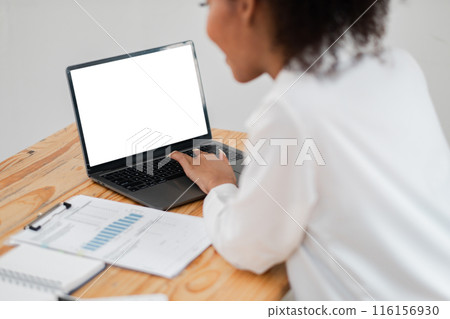 Focused woman using laptop at desk with business documents and charts, showcasing a professional work environment. 116156930