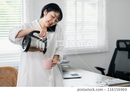 Young woman multitasking in a home office, pouring coffee while talking on the phone, bright and modern workspace. 116156969