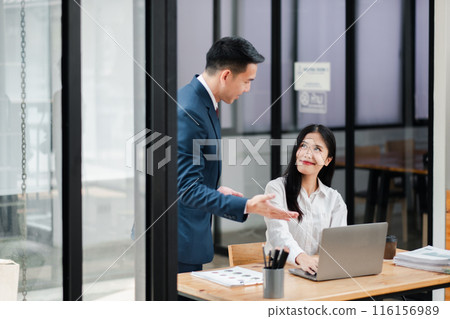 Businessman and businesswoman collaborating in a modern office, discussing work over a laptop, showcasing teamwork and professional interaction. 116156989